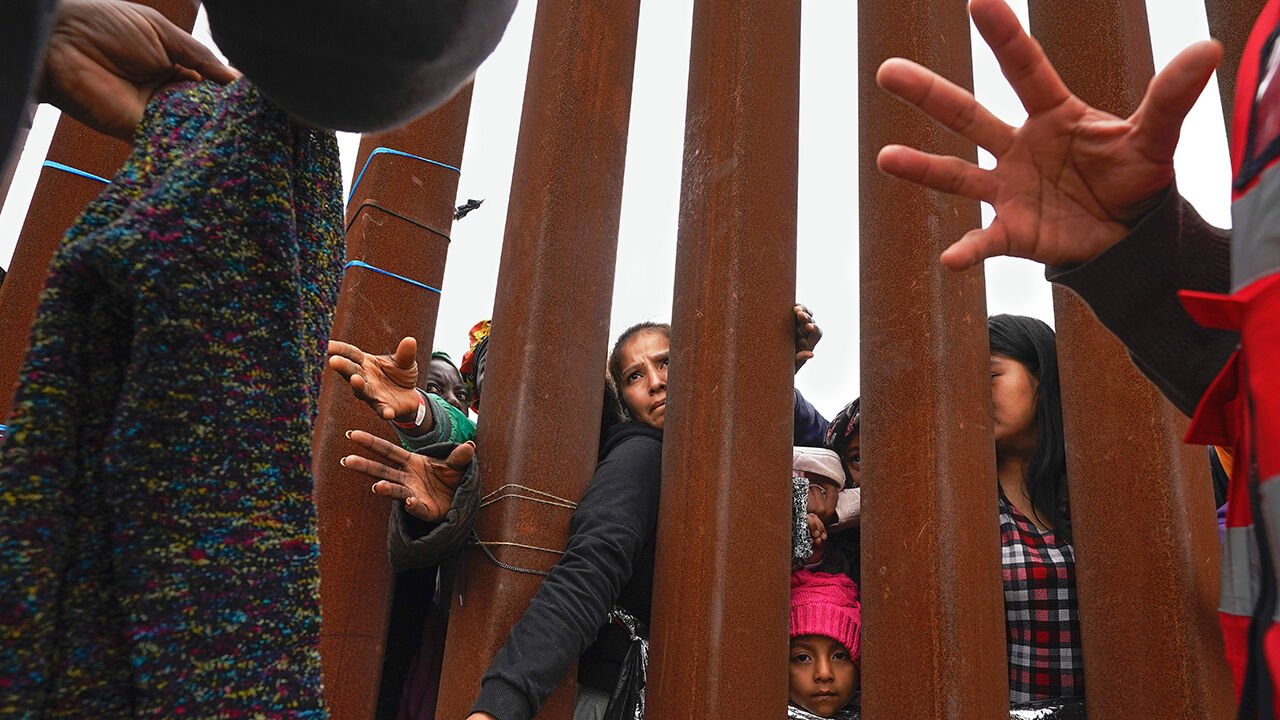 Migrants reach through a border wall in San Diego